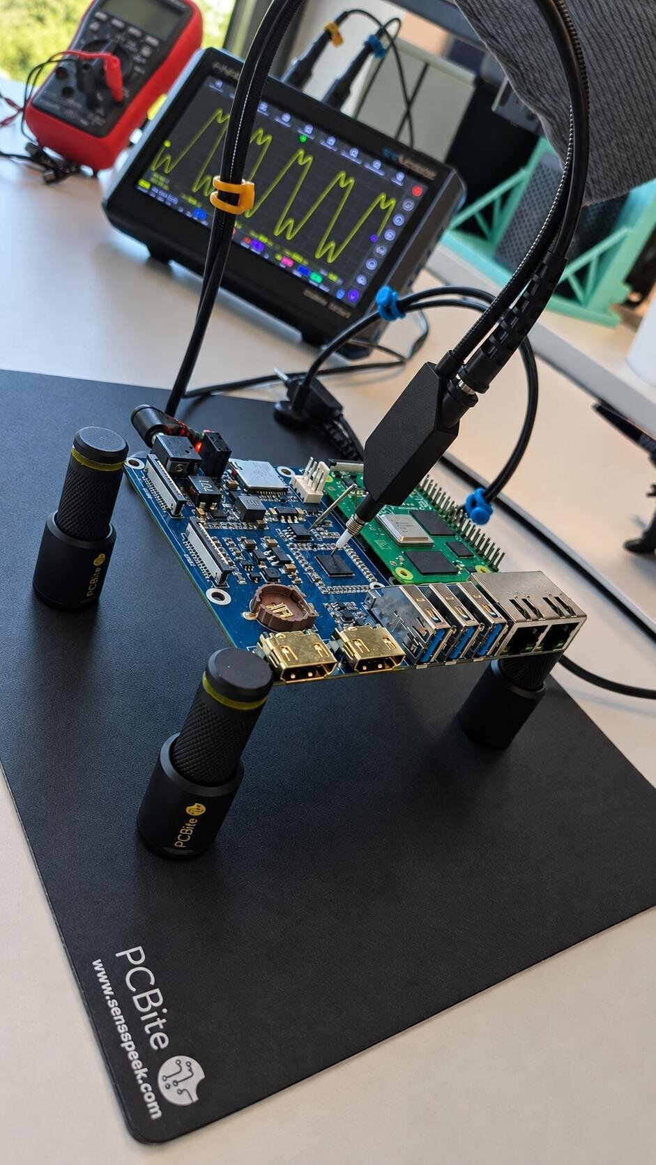 Circuit board under test probes, with an oscilloscope behind it